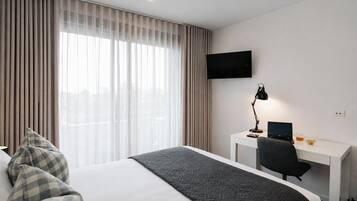 A hotel room with a bed, a desk with a computer, a TV, and a window with curtains.