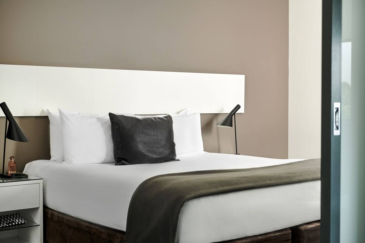 A neatly made bed with white and dark pillows, a green throw blanket, and a bedside table with a lamp and a bottle.