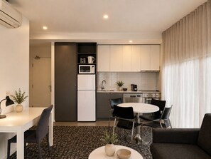 A modern kitchen with white cabinets, a built-in microwave, and a coffee machine. There is a dining area with a round table and chairs. - Punthill Oakleigh (Hughesdale)