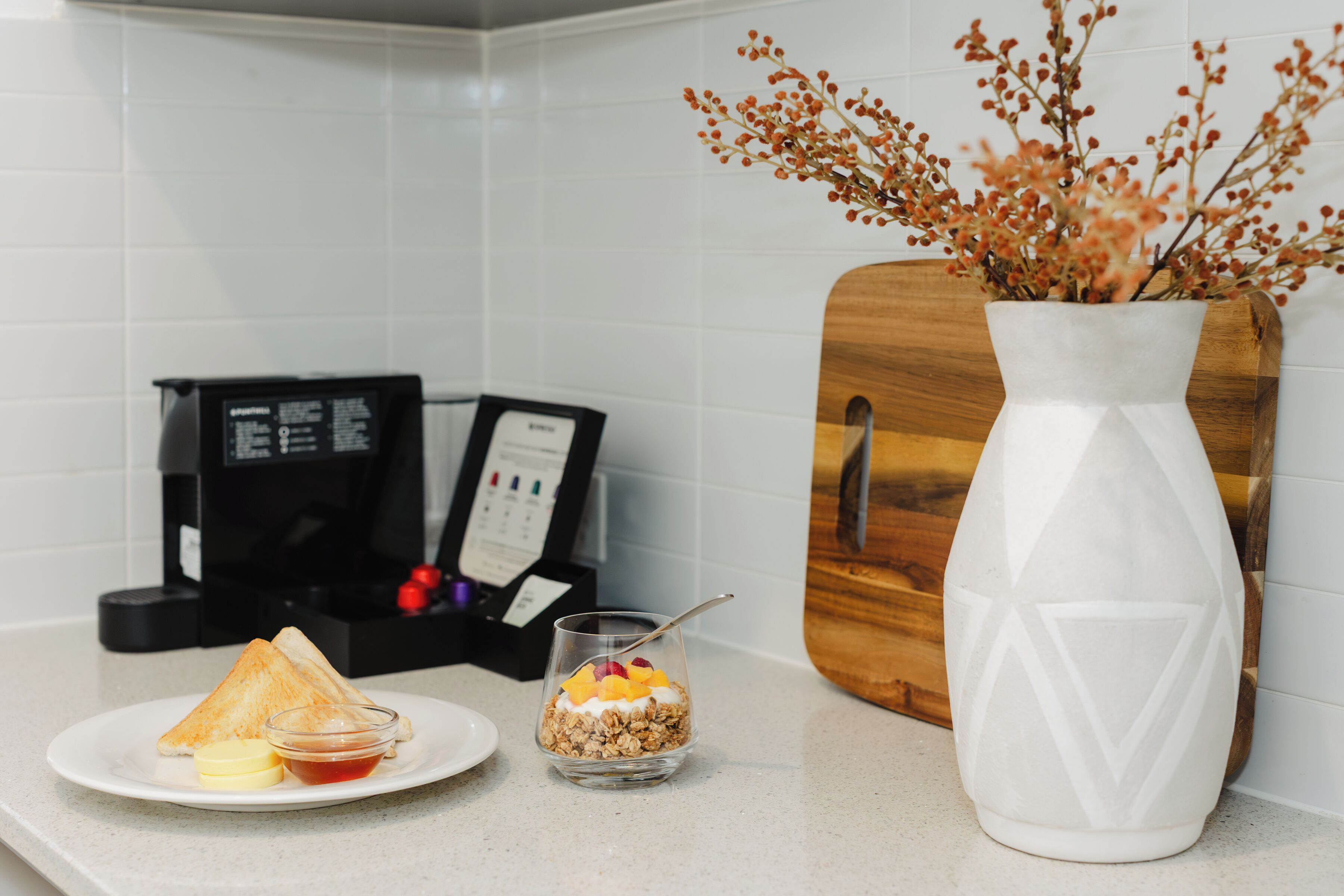 A breakfast setting with toast, jam, and a fruit bowl on a plate, a small bowl of cereal with fruit, and a white vase with dried flowers on a kitchen counter.