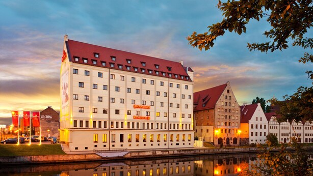 A large hotel with multiple floors and a red roof, situated by a calm river reflecting the building. - Qubus Hotel Gdansk (Gdansk)