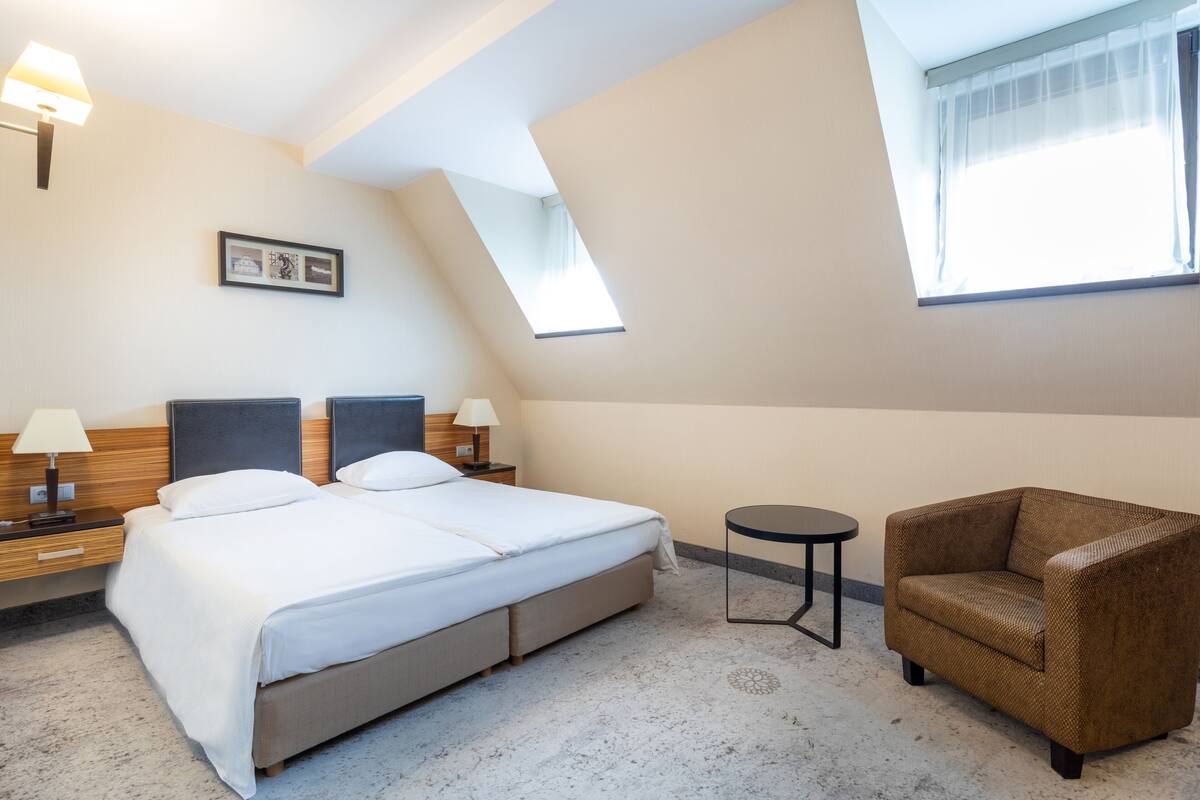 A hotel room with a large bed, two bedside tables with lamps, a small round table, a brown armchair, and a picture on the wall.