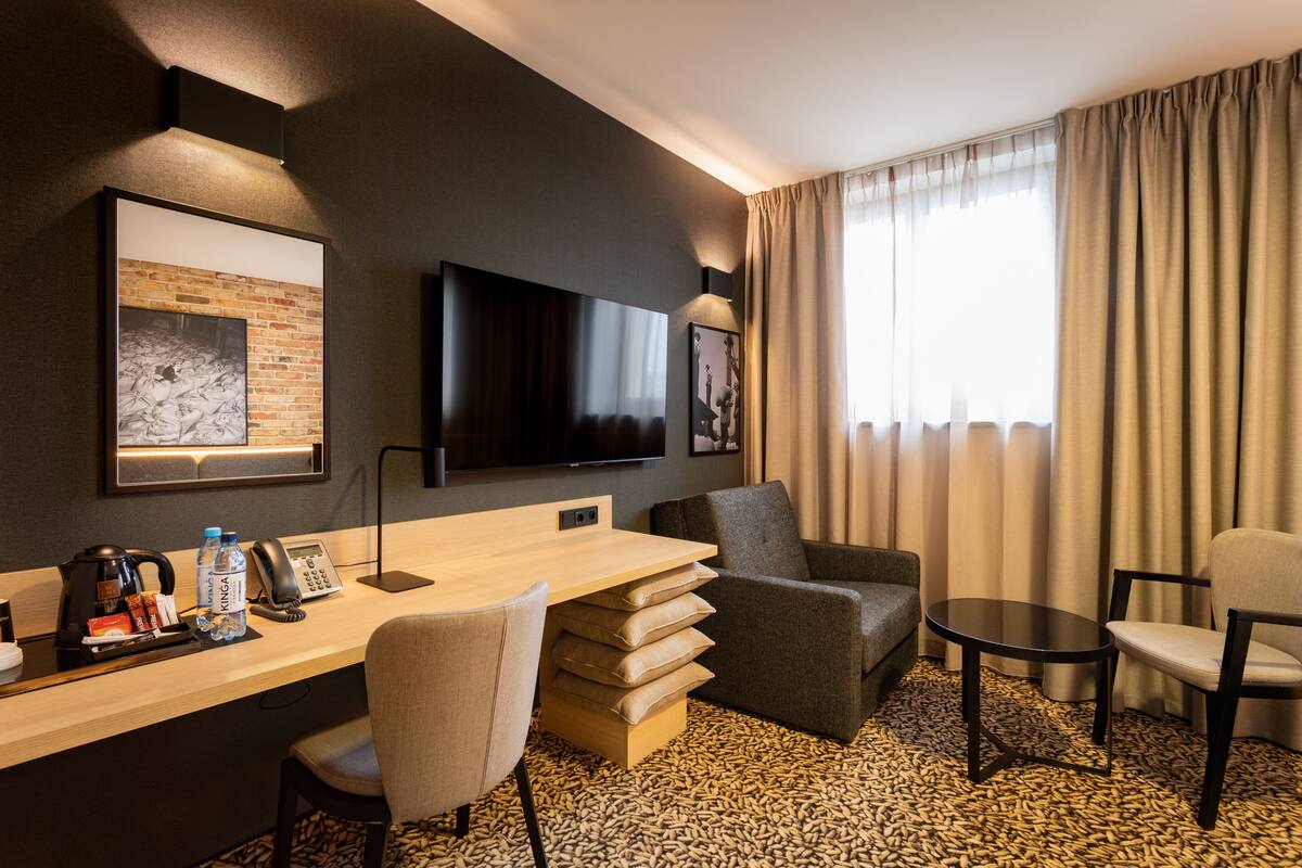 A hotel room with a desk, two chairs, a TV, and a large window with curtains.