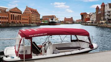 A boat with a red canopy and a website URL on the side, docked near a historic building with a large ship in the background.