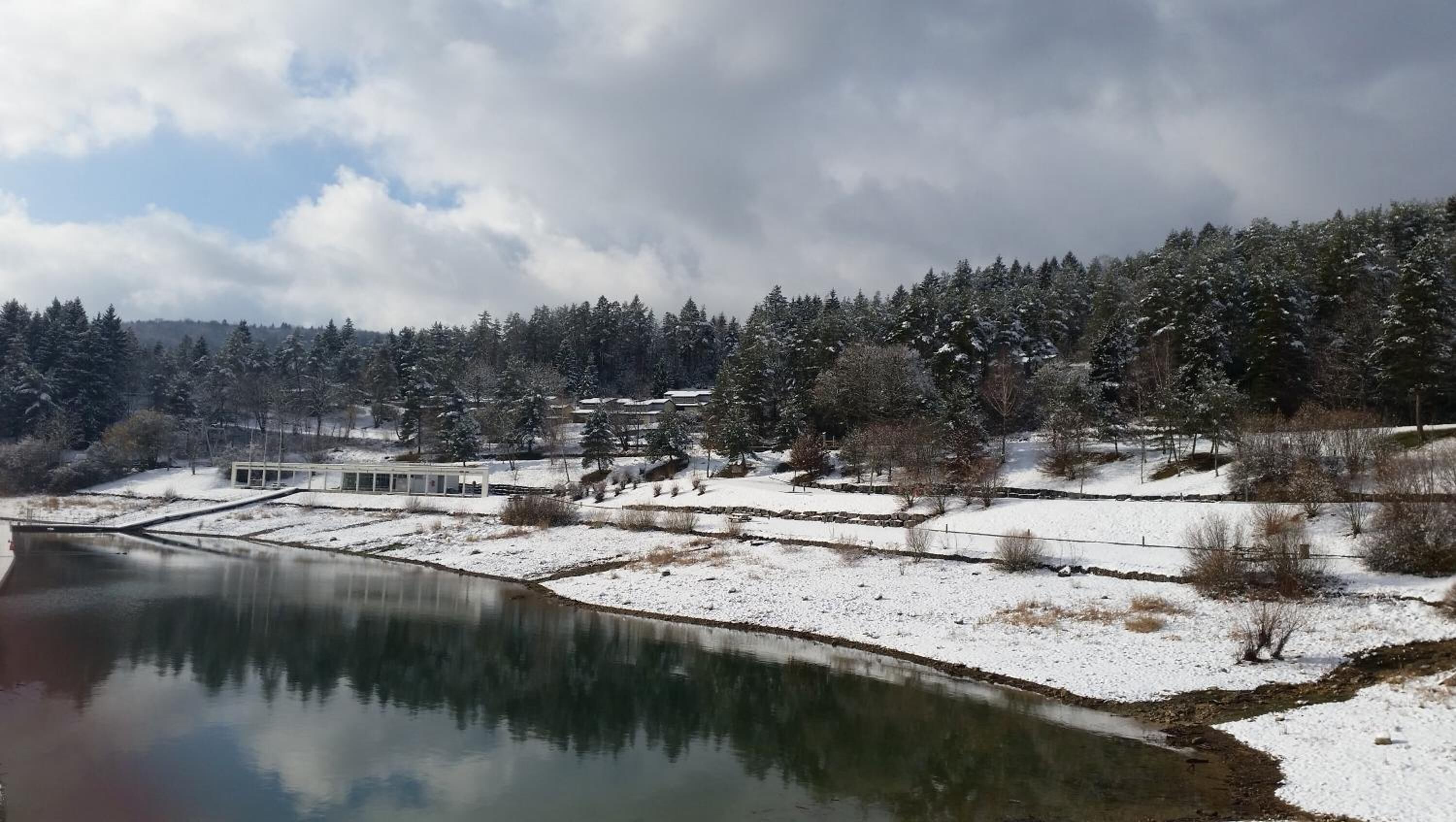 A serene lakeside scene with snow-covered ground, a small building, and dense trees in the background.