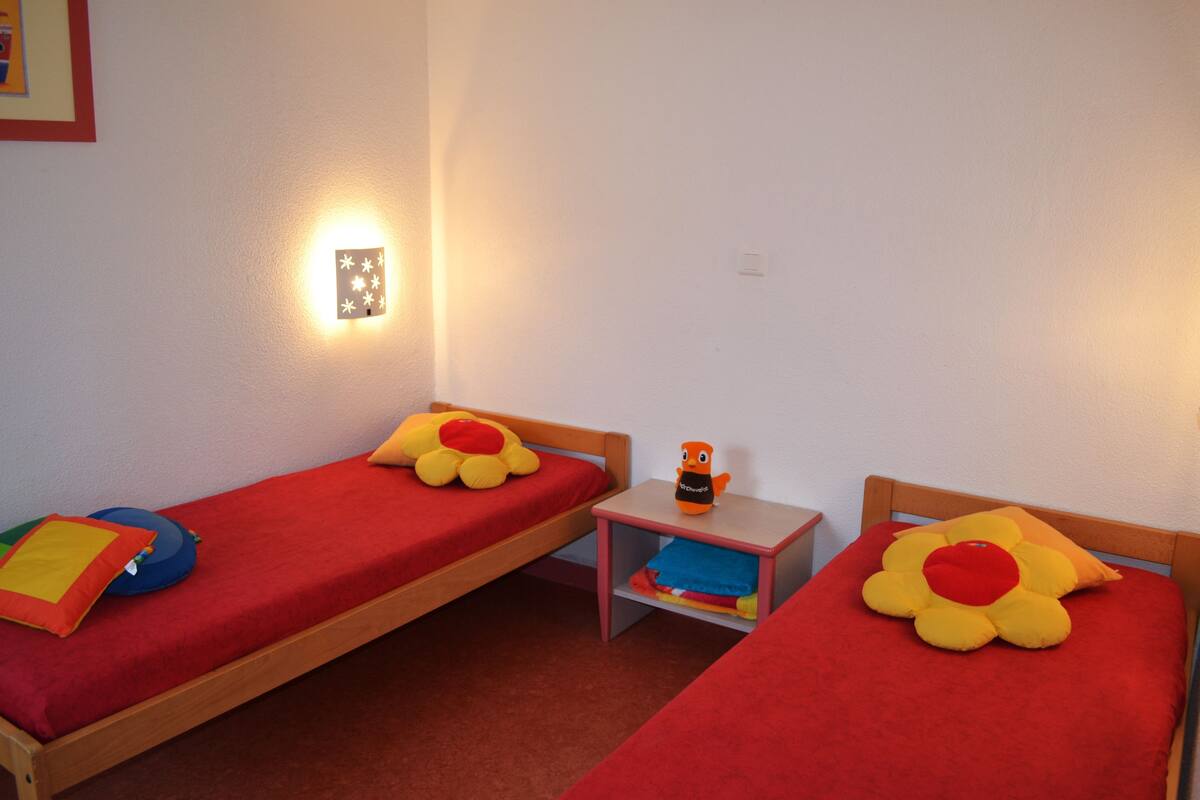 Una habitación con dos camas individuales, cada una con una colcha roja y un cojín con forma de flor. Entre las camas hay una mesita de noche con una lámpara.