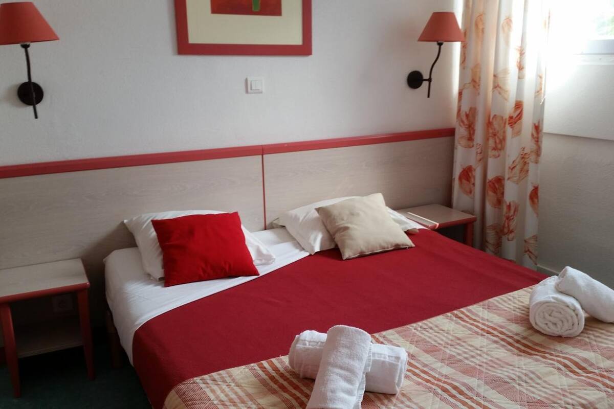 Habitación de hotel con cama, dos mesitas de noche, ropa de cama roja y blanca, dos lámparas de pared, un cuadro en la pared y una ventana con cortinas.