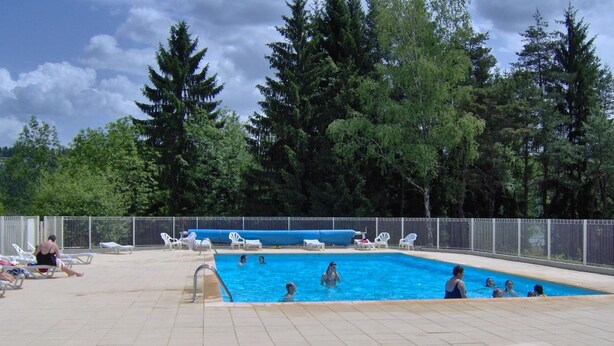 Ein Schwimmbecken mit Personen im Wasser und am Beckenrand, umgeben von Bäumen und einem eingezäunten Bereich mit Liegestühlen.