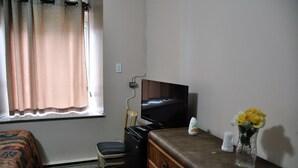 Quarto de hotel com cama, escrivaninha com computador, cadeira, televisão e um vaso com flores.