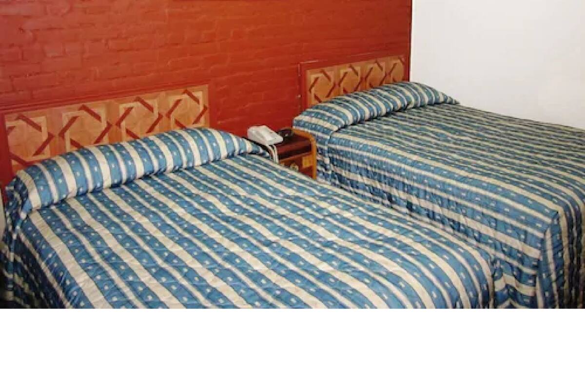 A room with two beds, each with striped bedding, a wooden headboard, and a nightstand with a phone. A painting hangs on the red brick wall above the beds.