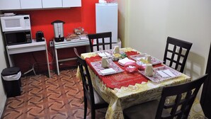 Uma cozinha compacta com micro-ondas, cafeteira e frigorífico. Uma mesa de jantar com cadeiras está disposta para as refeições.