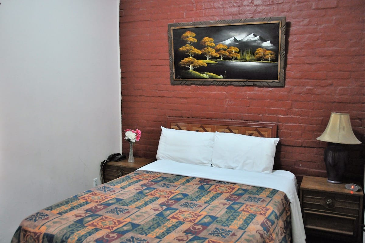 A room with a bed, a nightstand, a lamp, a picture on the wall, and a vase with flowers.