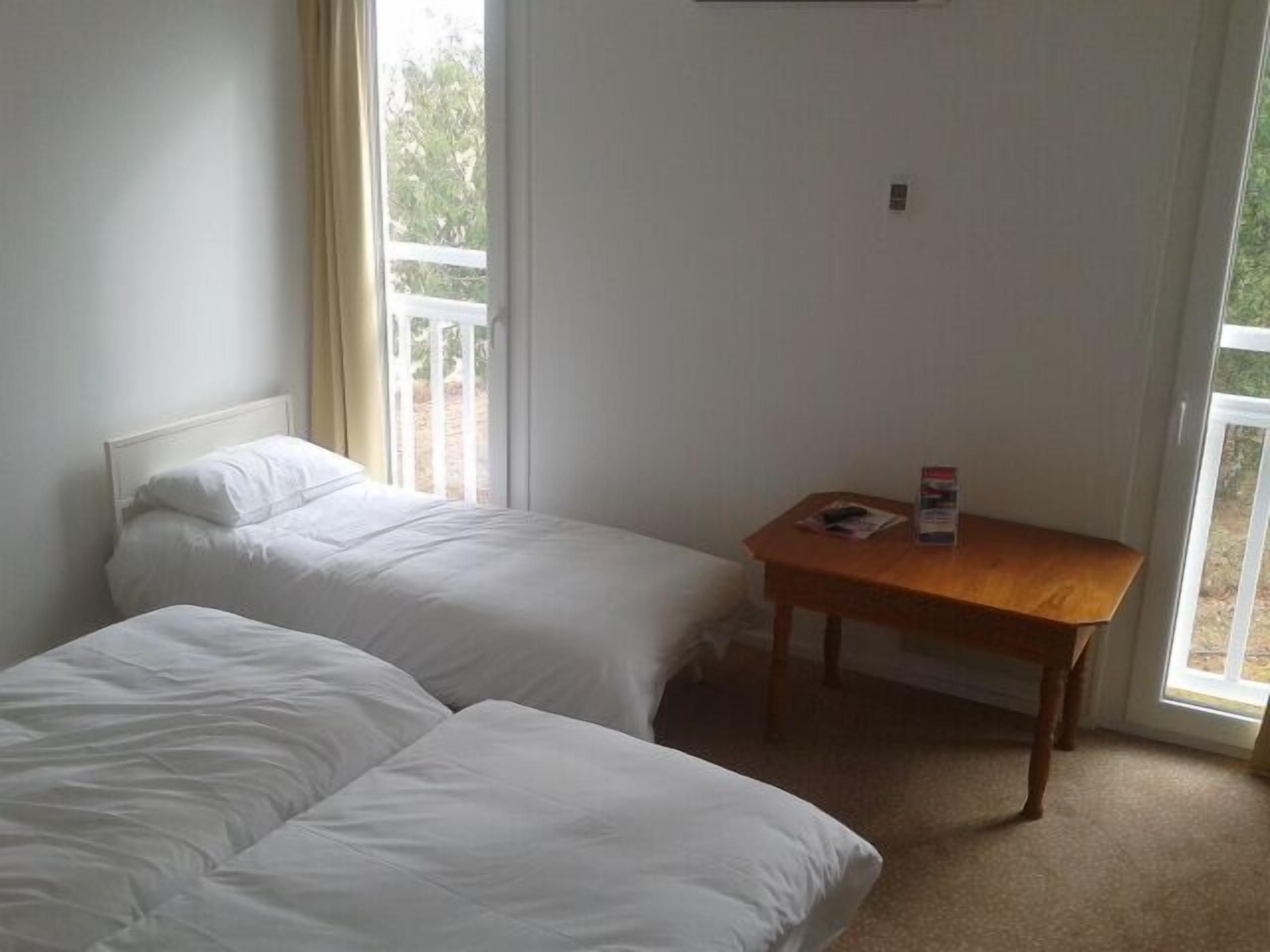 A room with two single beds, a wooden table, and a window with a view of greenery.