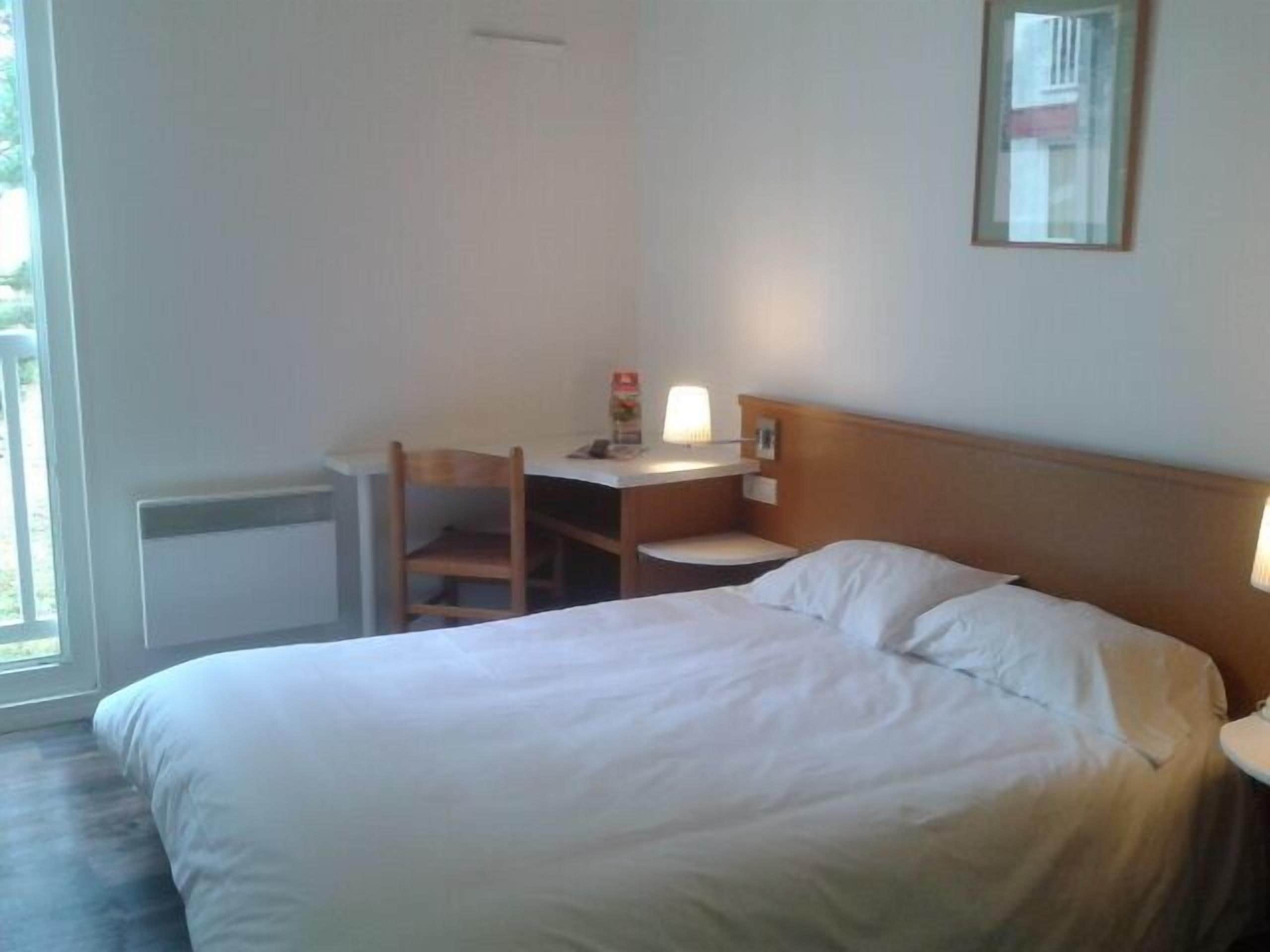 A hotel room with a bed, a desk with a chair, a lamp, and a window.
