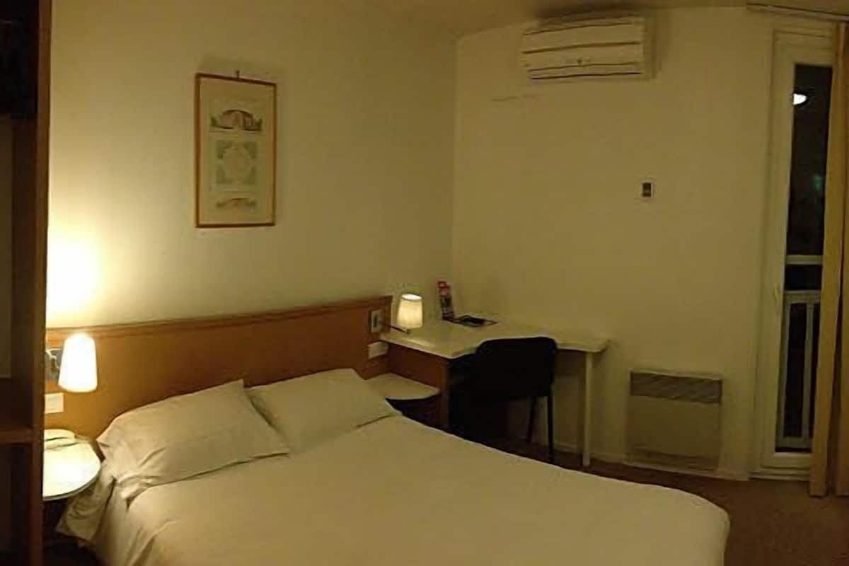 A hotel room with a bed, a desk, a chair, a lamp, and an air conditioning unit.