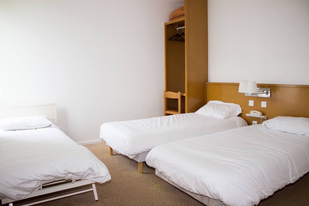 A hotel room with two single beds, a wooden wardrobe, a chair, and a bedside table with a lamp.