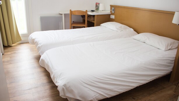 A hotel room with two single beds, a wooden desk, a chair, a lamp, and a window with curtains.