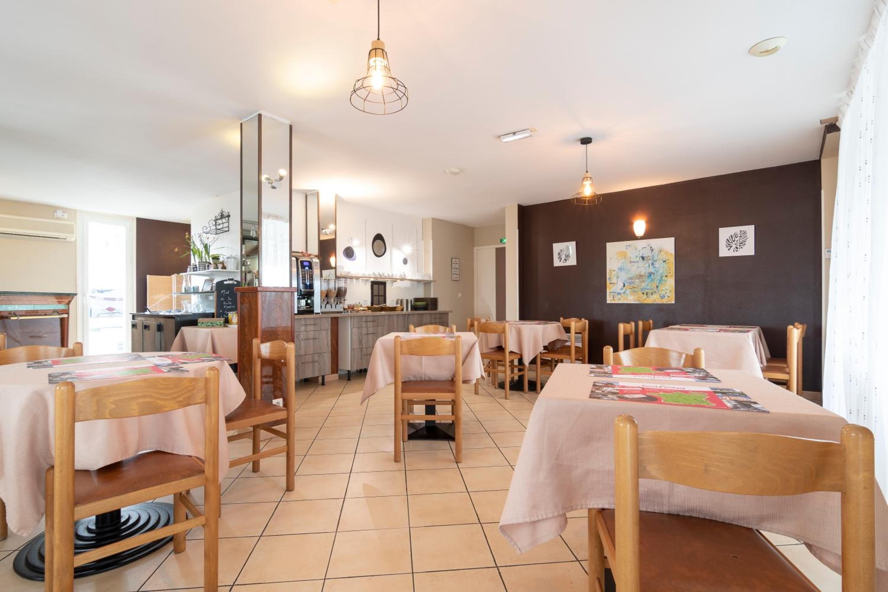 A dining area with wooden tables and chairs, a kitchen in the background, and artwork on the walls.