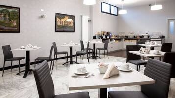 A modern dining area with marble flooring, black upholstered chairs, and white tables. There are framed artworks on the walls and a kitchen area in the background.