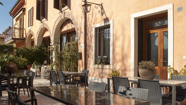 Un café en plein air, avec des tables et des chaises, entouré de plantes et bordé par un bâtiment doté d’arcades et d’une porte en bois.