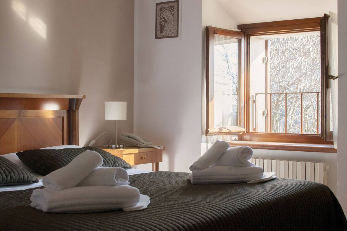 Une chambre d’hôtel avec deux lits, une tête de lit en bois, une table de chevet avec une lampe, un téléphone et une fenêtre donnant sur des arbres.