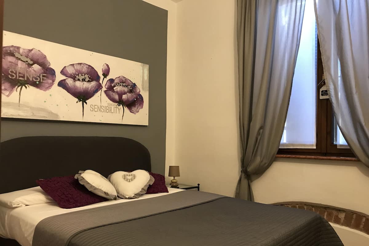 Un lit bien fait, avec une couvre-lit grise et des oreillers blancs, une tête de lit sombre et un tableau accroché au mur représentant des fleurs violettes, sur lequel figure le mot « SENSIBILITÉ ».