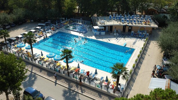 A large outdoor swimming pool with multiple lanes, surrounded by lounge chairs and umbrellas, and a building with a roof overlooking the pool. - Hotel Beau Soleil (Cesenatico)