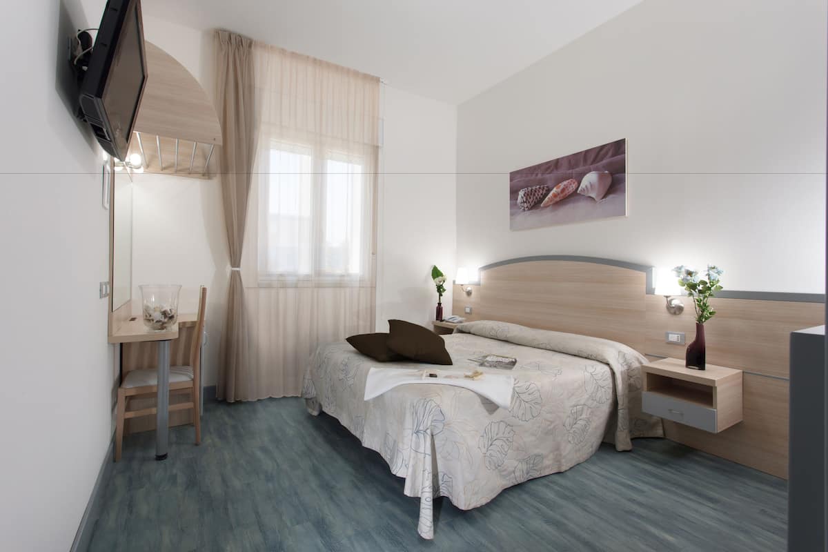 Ein Hotelzimmer mit Bett, Nachttisch, Sessel, Fernseher und einem Fenster mit Vorhängen.