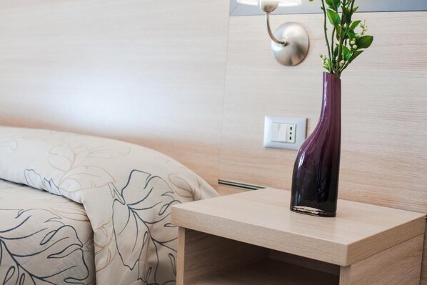 A bed with a floral-patterned duvet, a bedside table with a vase of green plants, and a wall-mounted light fixture. - Hotel Beau Soleil (Cesenatico)