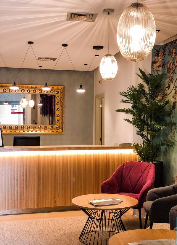 Un área de recepción con un mostrador de madera, un sillón rojo, un sillón gris y una planta grande en maceta.
