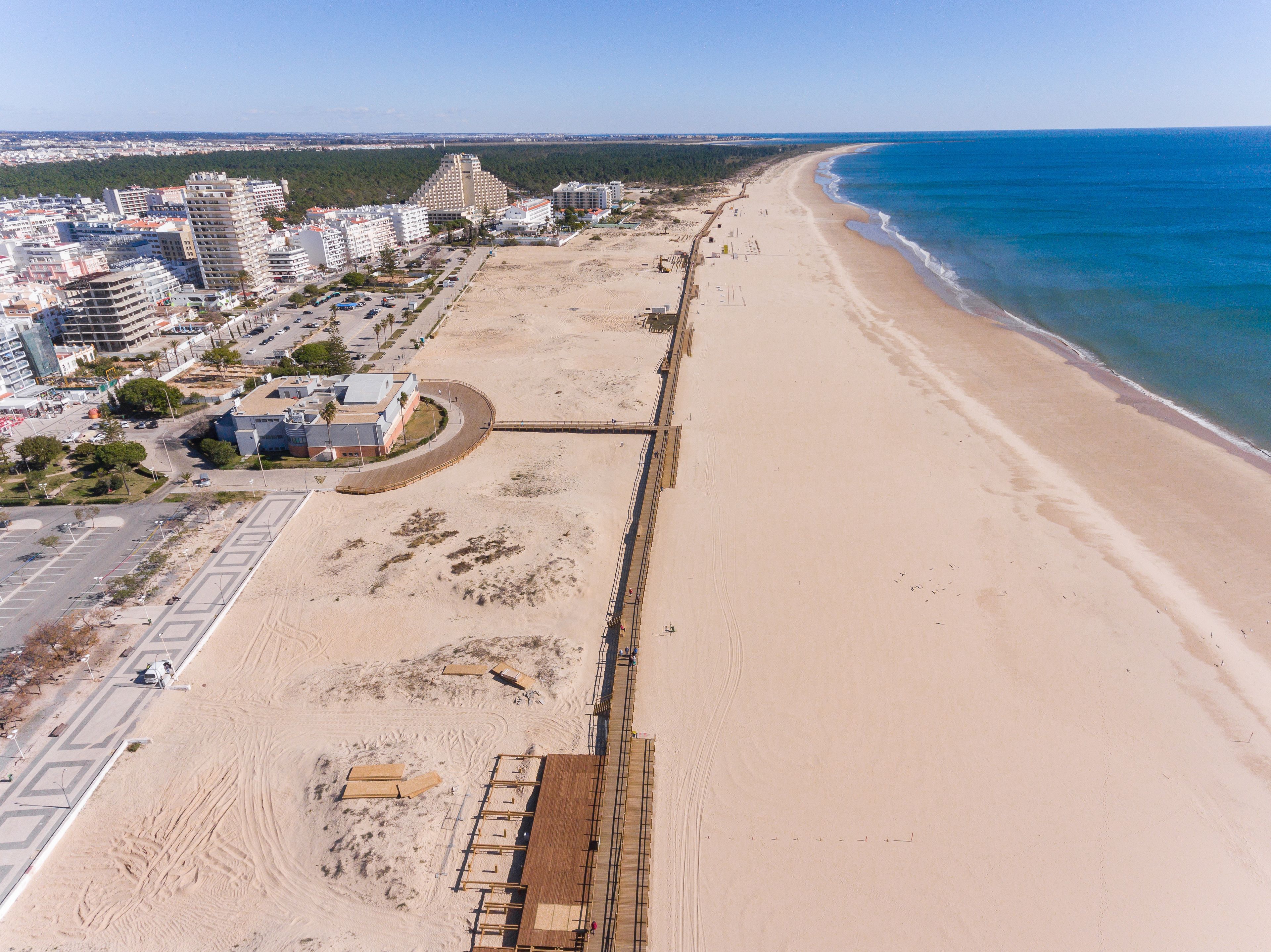 Vista aérea de una zona costera con una playa de arena, un paseo marítimo y una hilera de edificios a lo largo de la orilla.