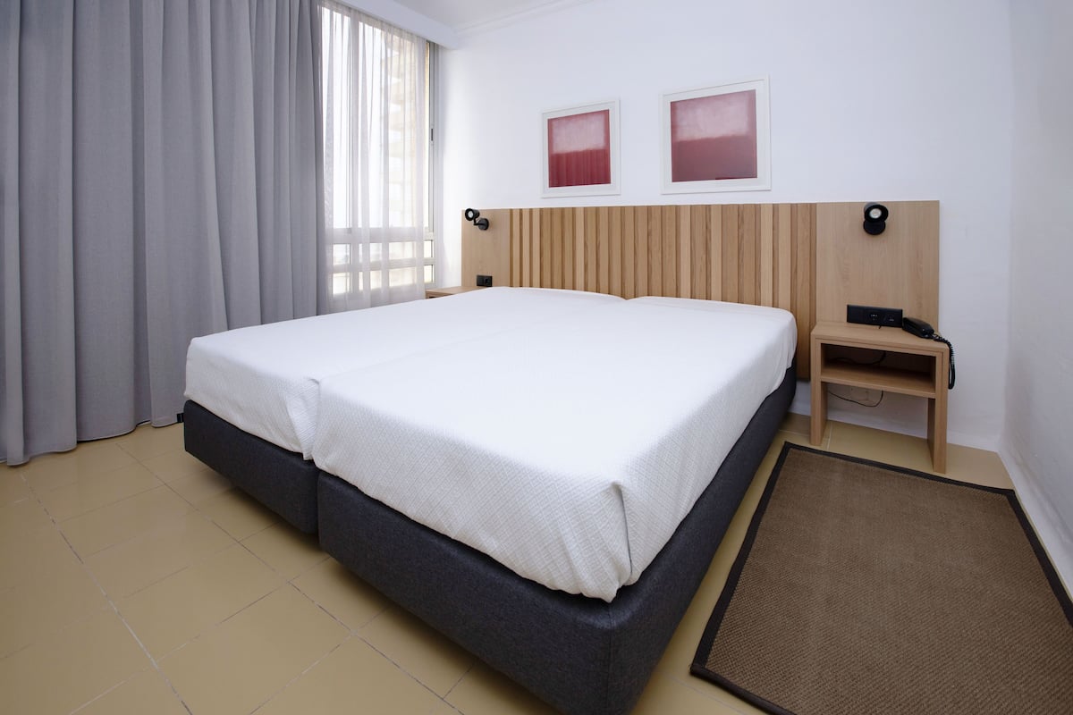 Une chambre d’hôtel avec un grand lit, une table de chevet avec un téléphone, une lampe fixée au mur et deux tableaux encadrés.