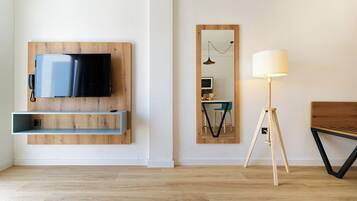 A modern living room with a mounted TV, wooden shelves, a standing lamp, and a mirrored wall.