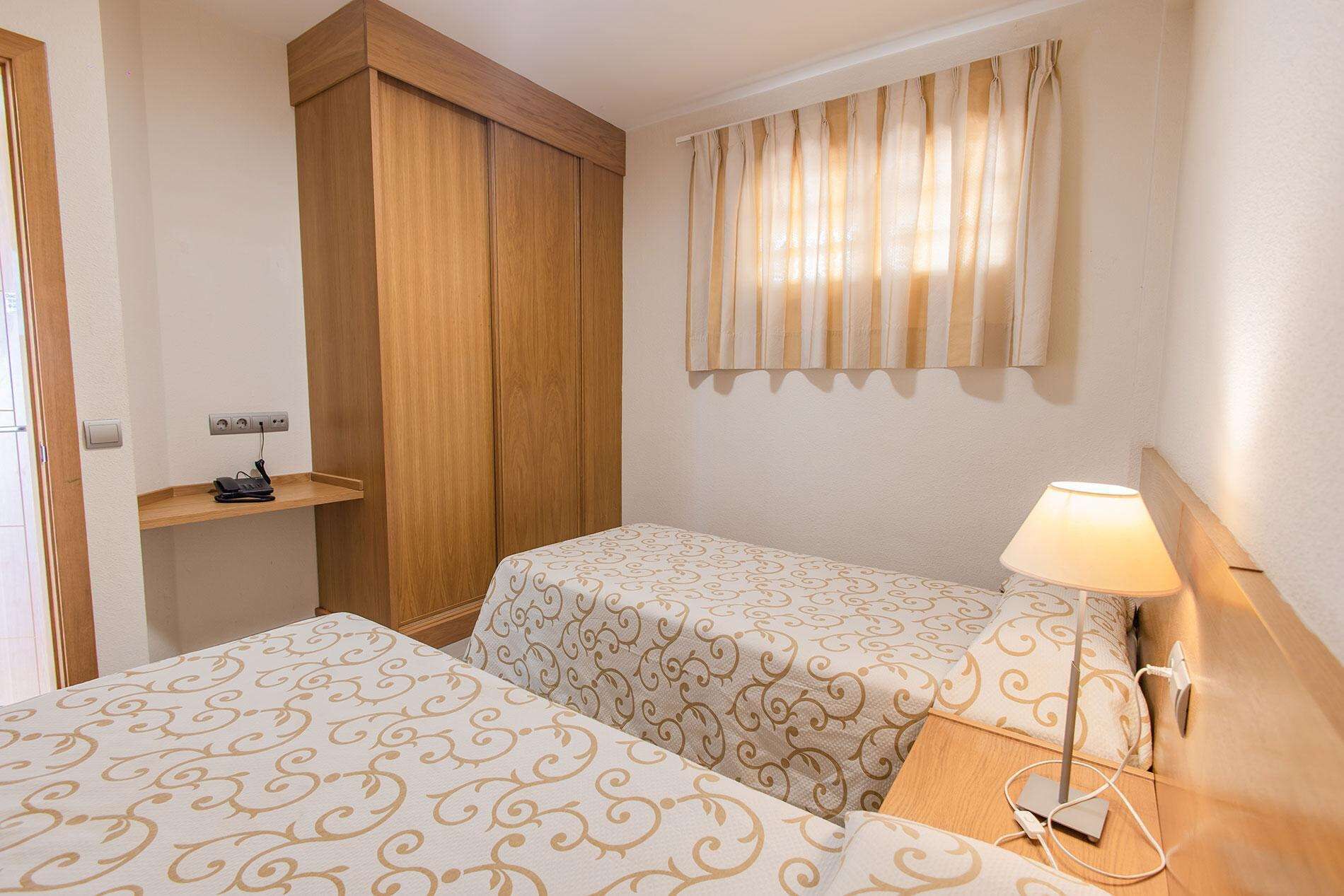 A hotel room with two beds, a wooden wardrobe, a bedside table with a lamp, and a window with curtains.