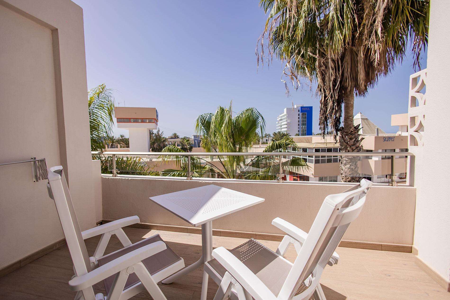 A balcony with two white chairs and a table, overlooking a cityscape with palm trees and buildings.