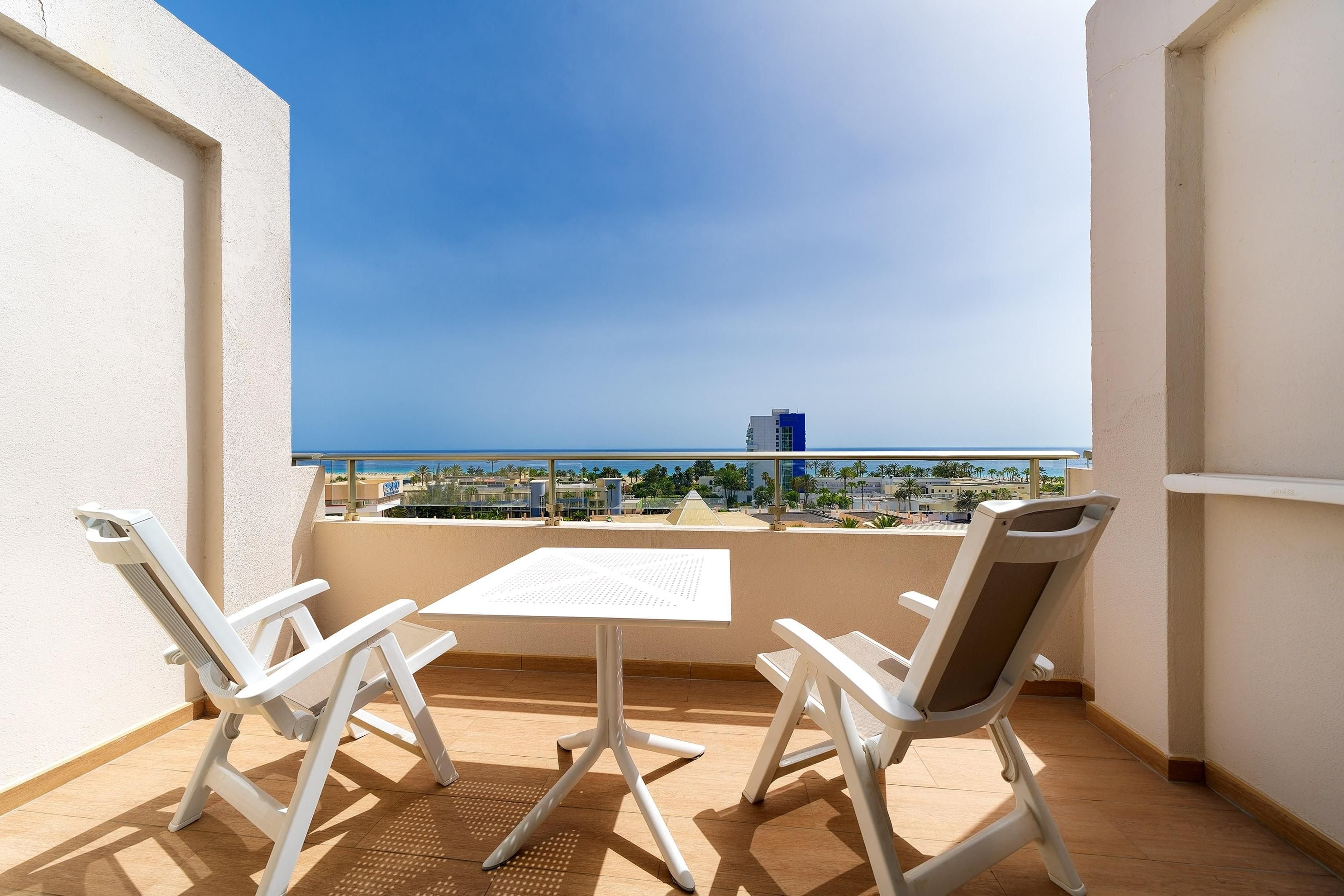 Un balcon avec deux chaises blanches et une table, d’où l’on peut admirer un paysage urbain côtier sous un ciel dégagé.