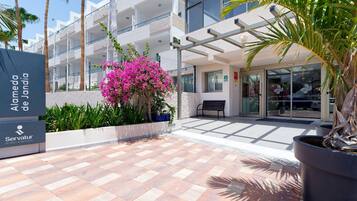 Entrada de un hotel con un sendero pavimentado, una banca y un letrero que dice 'Alameda de Jandía Servatur Hotels & Resorts'.