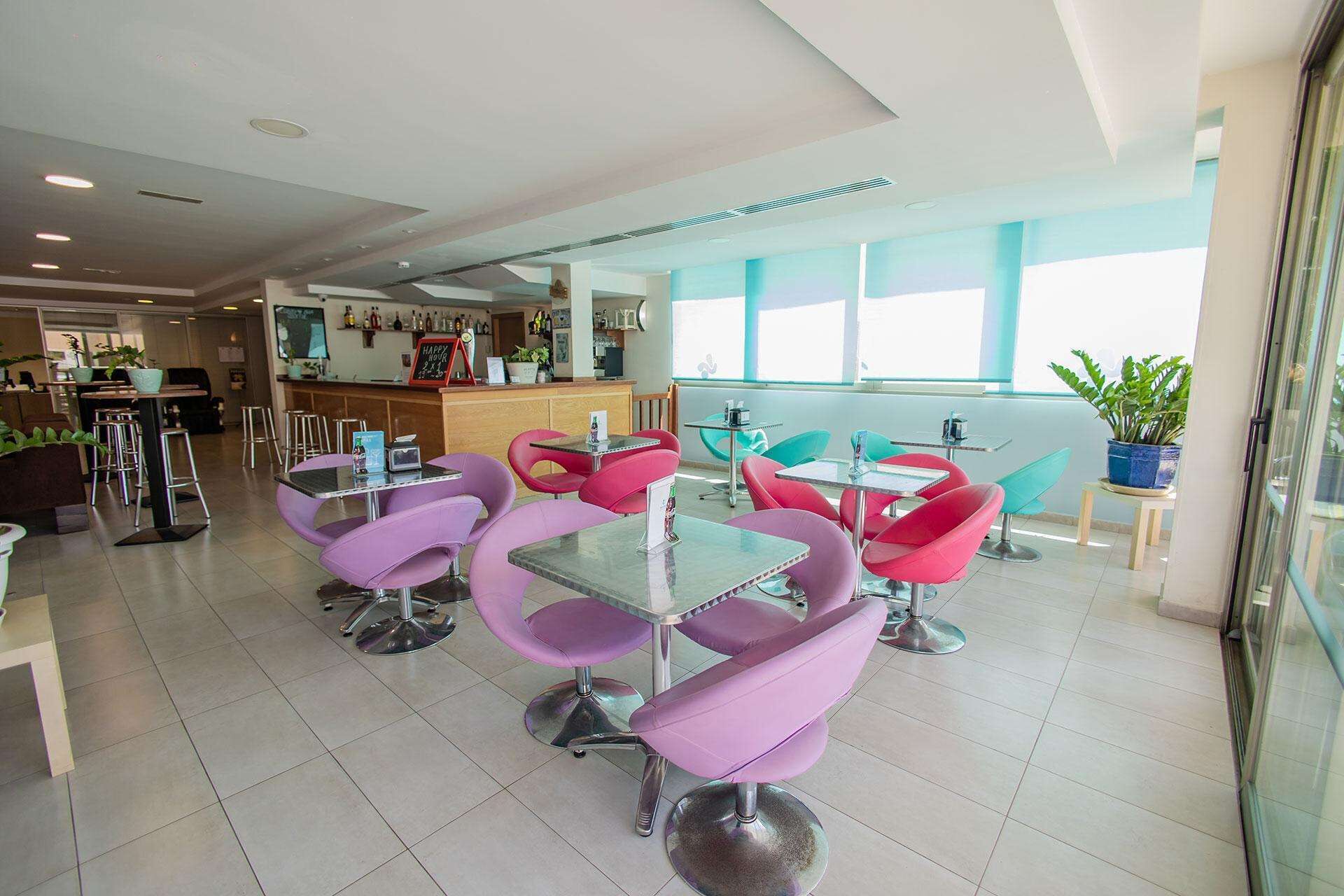 Un espace bar moderne avec des tabourets et un comptoir, un coin salon avec des tables et des chaises rondes, et une vue sur l’extérieur grâce à de grandes fenêtres.