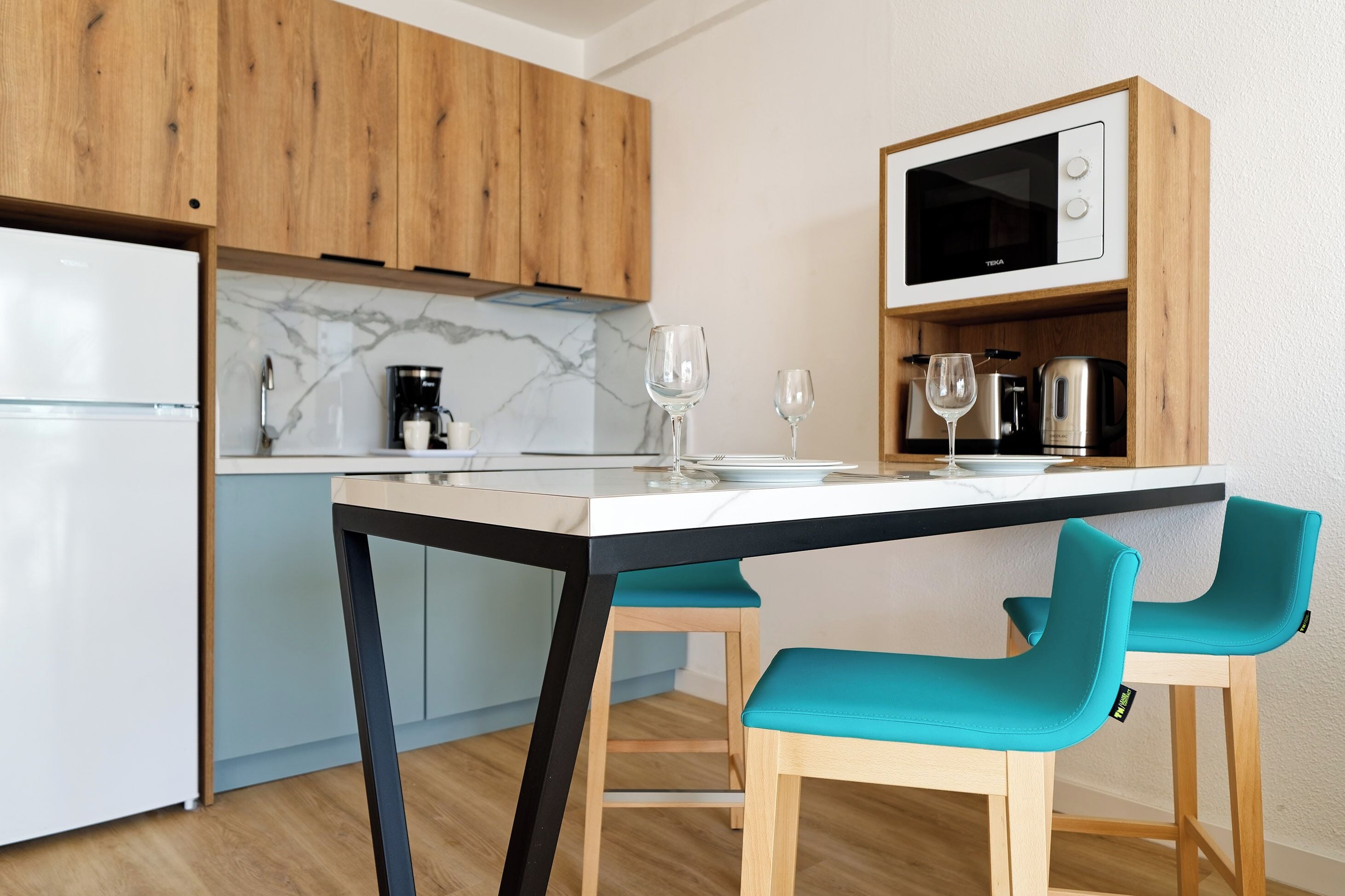 A modern kitchen with wooden cabinets, a white refrigerator, a black and white microwave, and two teal chairs.