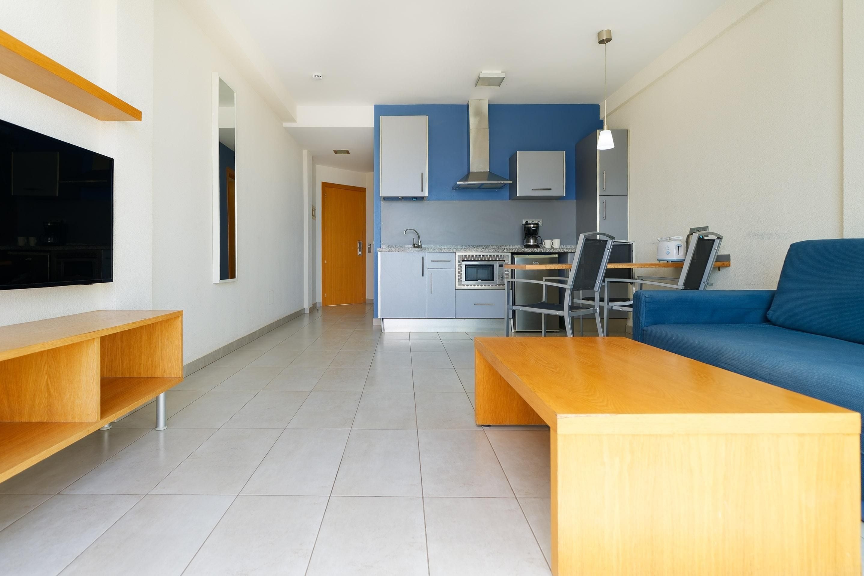 Un salon moderne avec un téléviseur, des meubles en bois, un canapé bleu et une cuisine en arrière-plan.