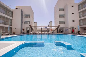 A swimming pool with a wooden deck, surrounded by white buildings with balconies. - Servatur Alameda de Jandía (Pajara)