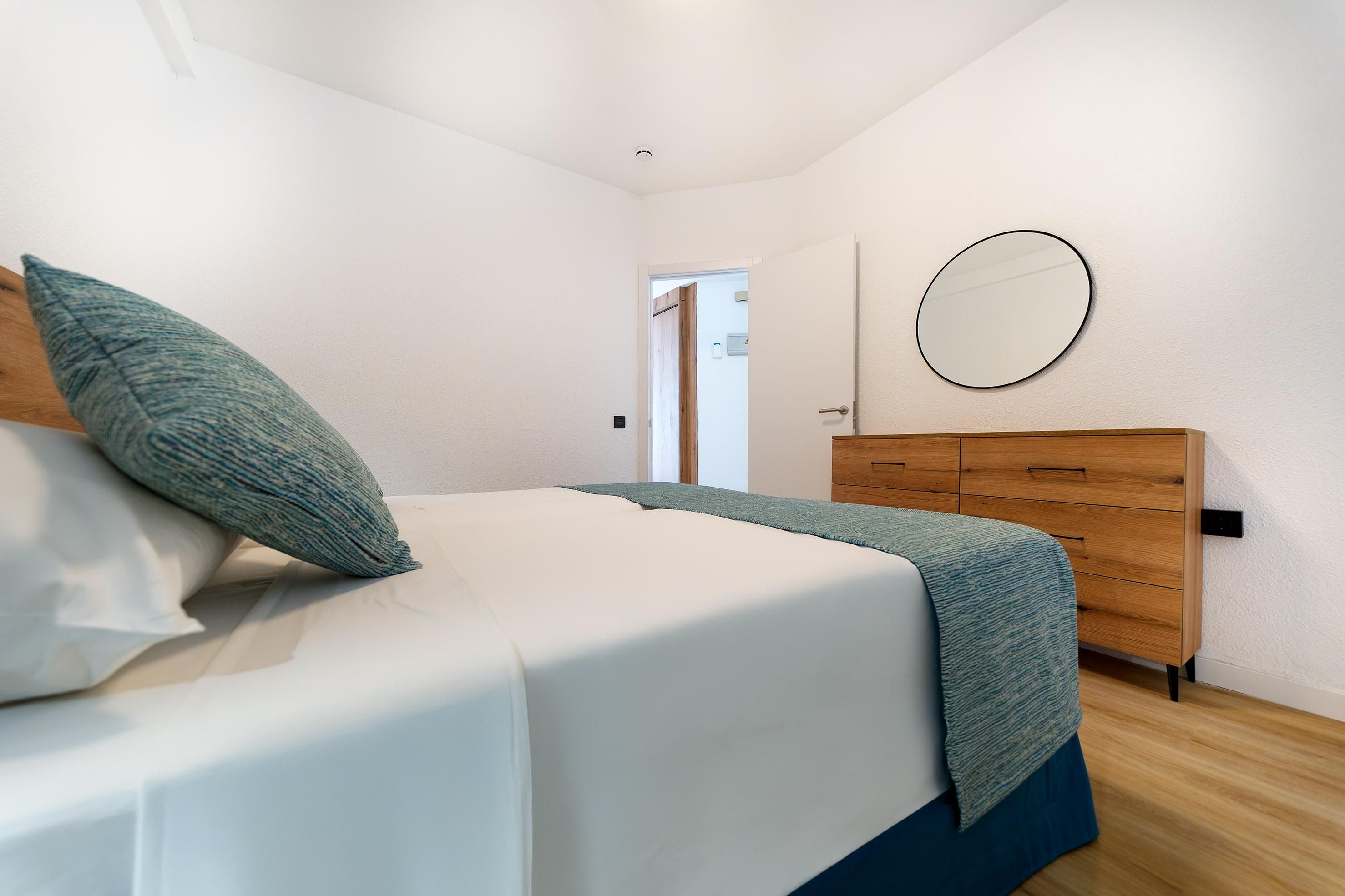 Un dormitorio con cama, cómoda de madera, espejo redondo y puerta que da a otra habitación.