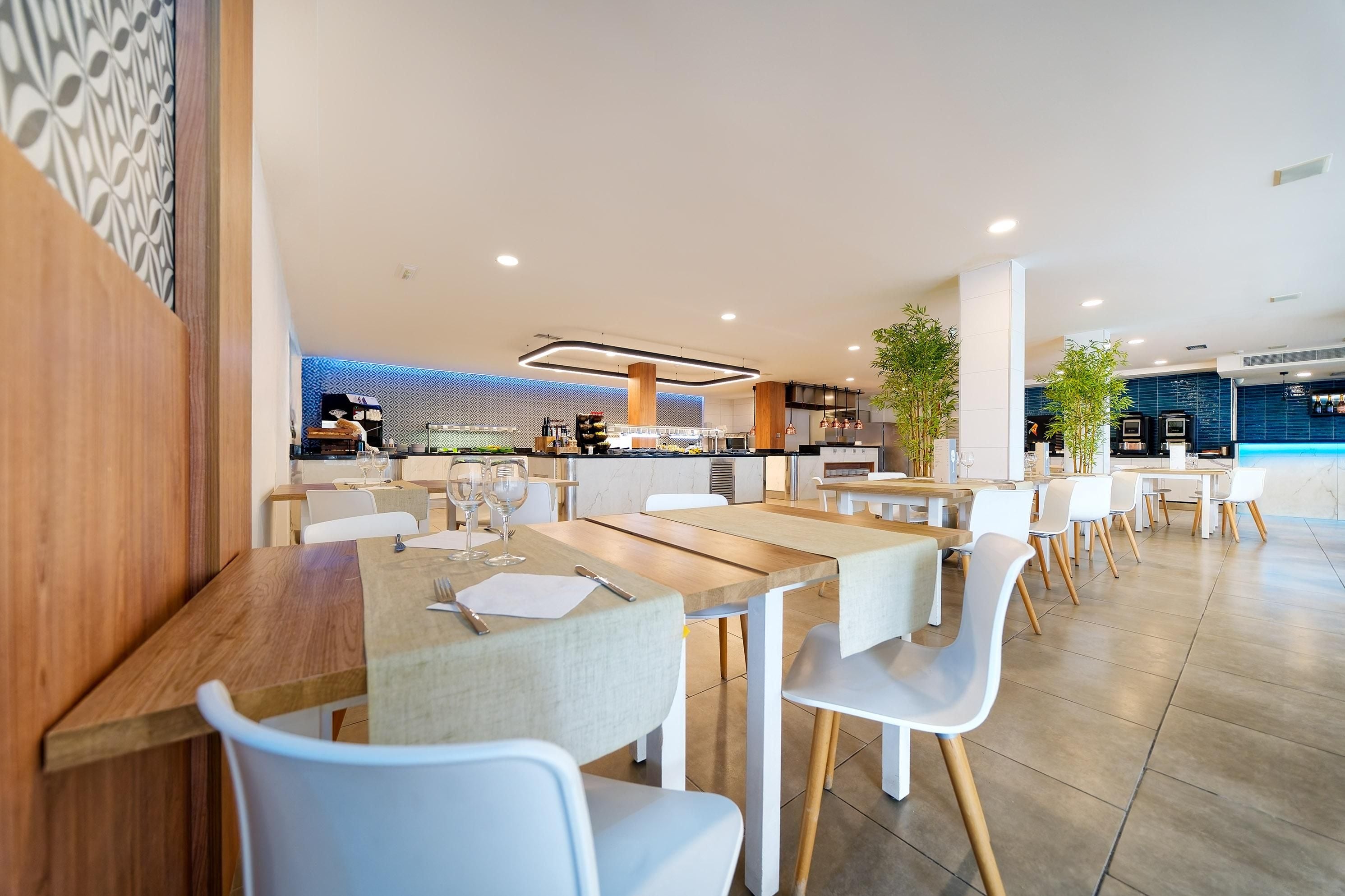 Un restaurante moderno con mesas de madera y sillas blancas, un área de cocina al fondo y una pared con diseño azulado.