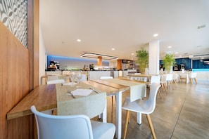 Un restaurante moderno con mesas de madera y sillas blancas, un área de cocina al fondo y una pared con diseño azulado.