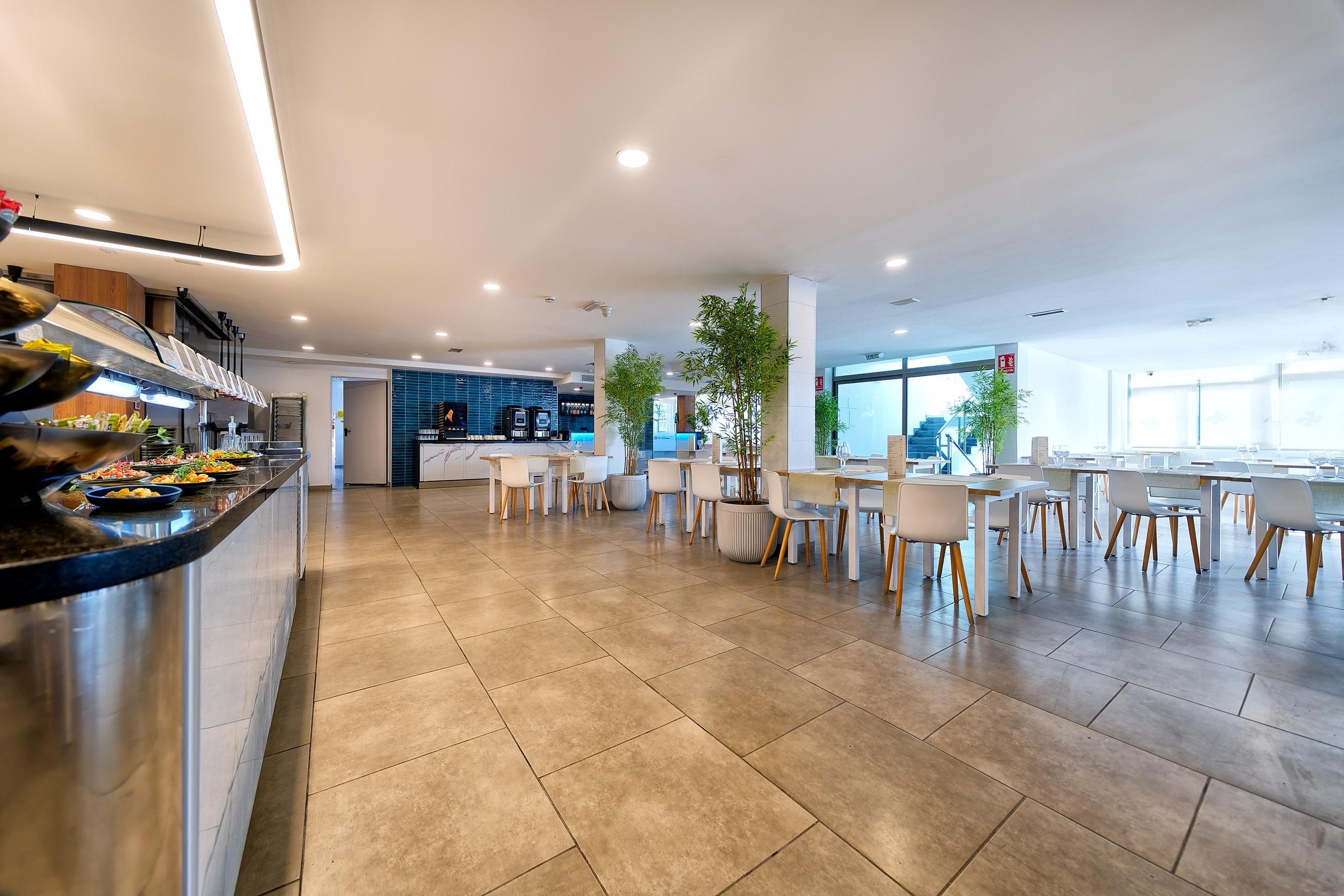 Una cafetería moderna con una zona de bufé, mesas y sillas, y una cocina al fondo.