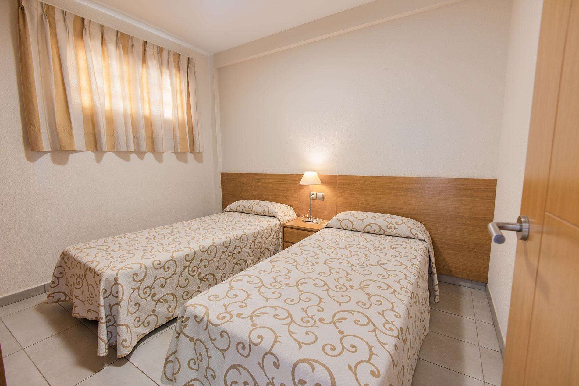 A room with two single beds, a wooden headboard, a bedside lamp, and a window with curtains.