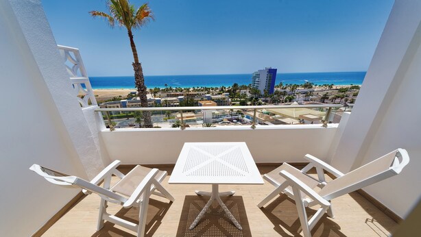 A balcony with a white table and chairs, overlooking a beach and ocean. - Servatur Alameda de Jandía (Pajara)
