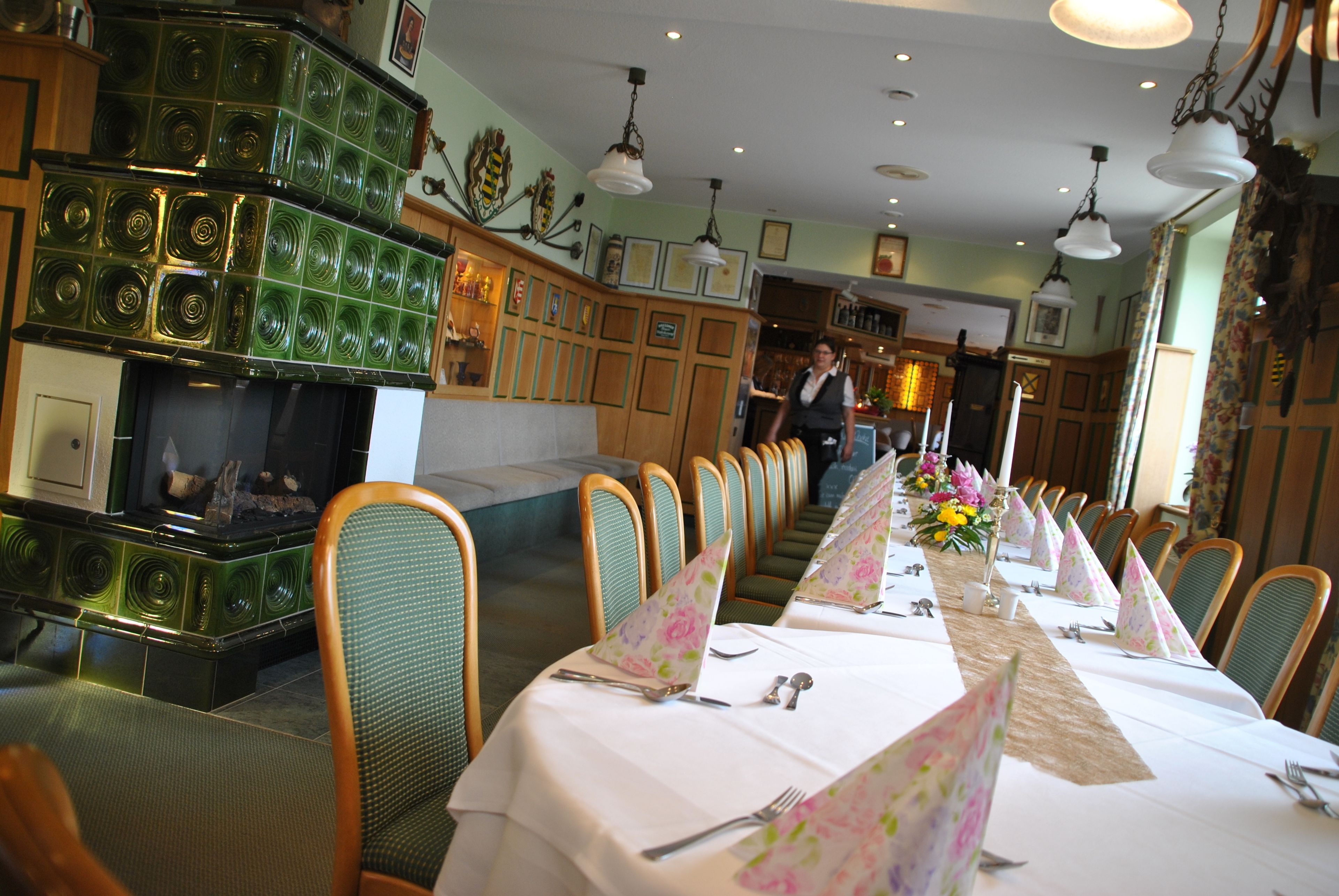 Restaurante com uma longa mesa de jantar posta para uma refeição, cadeiras de madeira e um bar com copos verdes.
