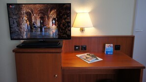 Habitación de hotel con una televisión instalada en un mueble de madera, una mesa con una revista, una lámpara y una pequeña señal en la pared.