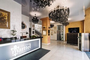 Un hall d’hôtel avec un comptoir d’accueil, une grande horloge et un escalier menant aux étages supérieurs.